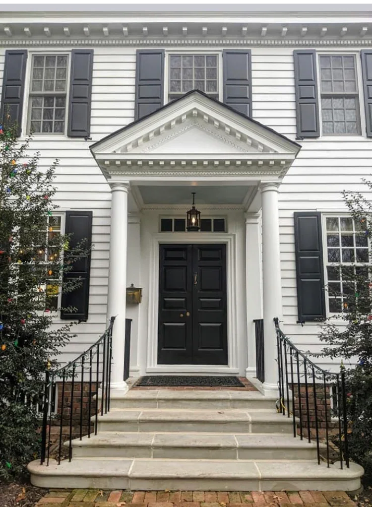White colonial home with dark shutters, painted portico with columns, black double doors, and detailed trim