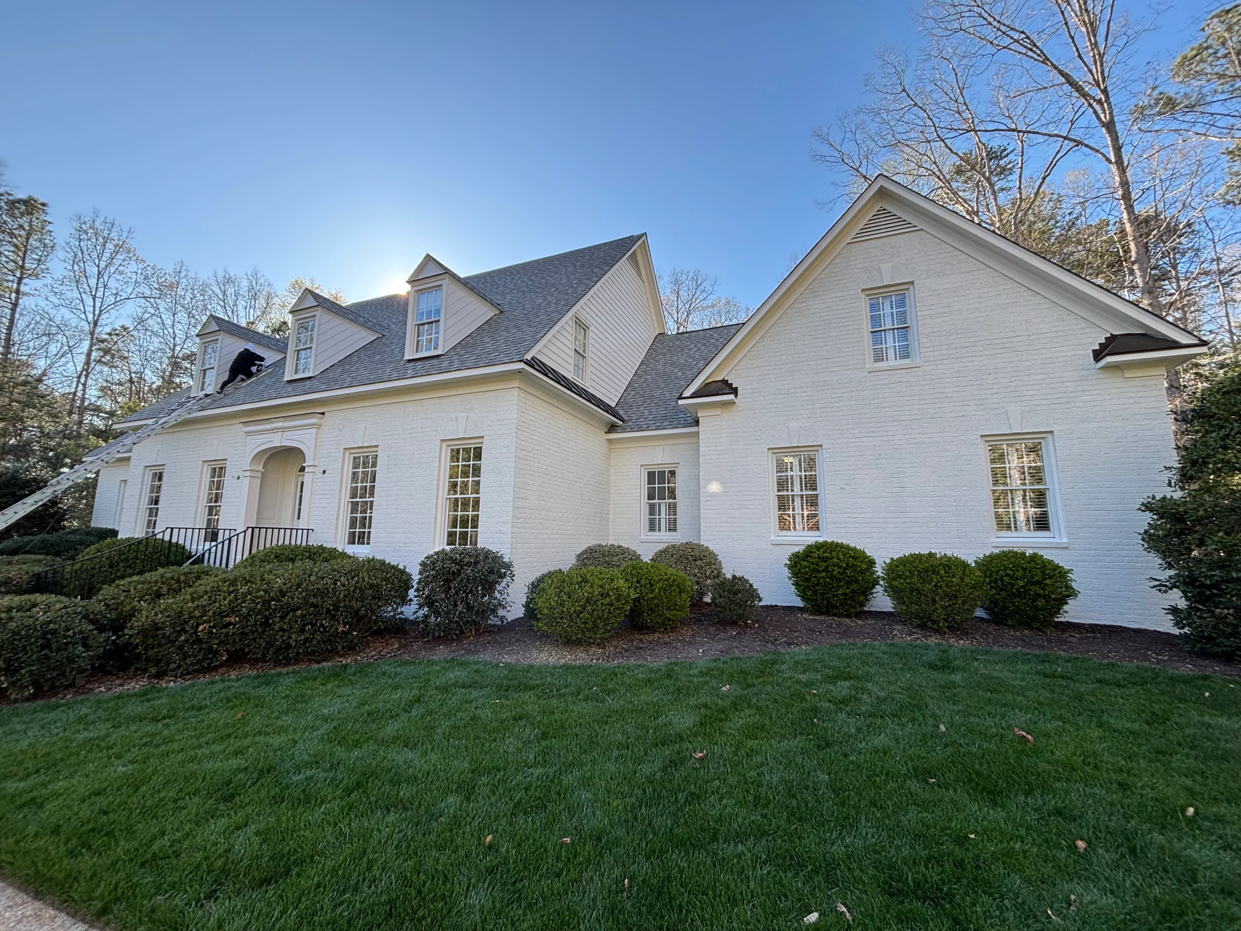 White brick home with dormers, dark slate roof, painted trim, and manicured landscaping